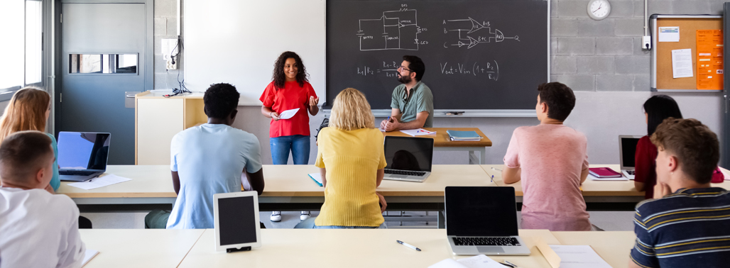 high school aged students sitting at desks and looking in the front of a room where a female student is presenting and a male student is sitting on the table next to her. Chalk board with circuit diagrams are on it in the background.