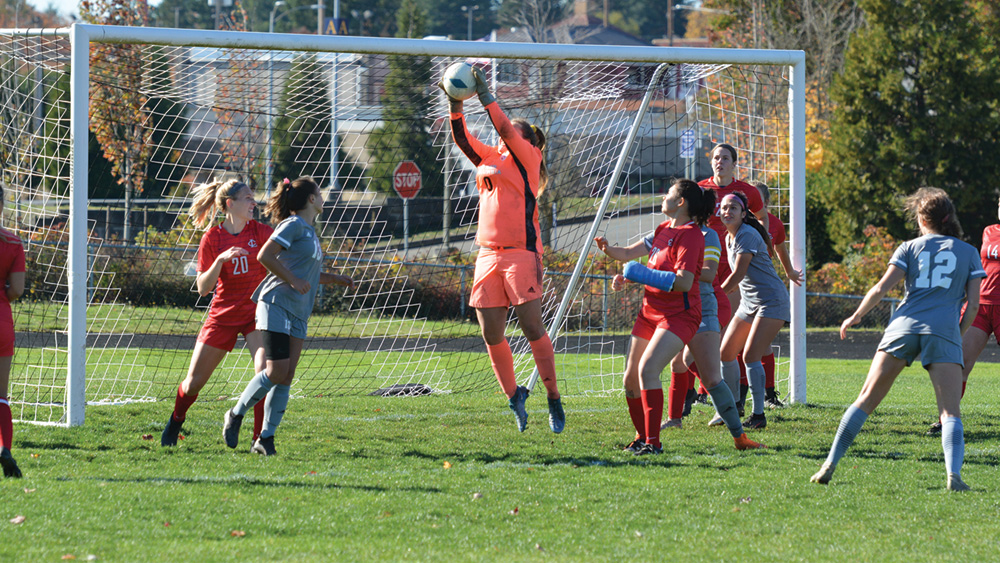 LCC women's soccer game, players in red and grey jersey's around goal as goal keeper in orange jersey jumps to catch the soccer ball.